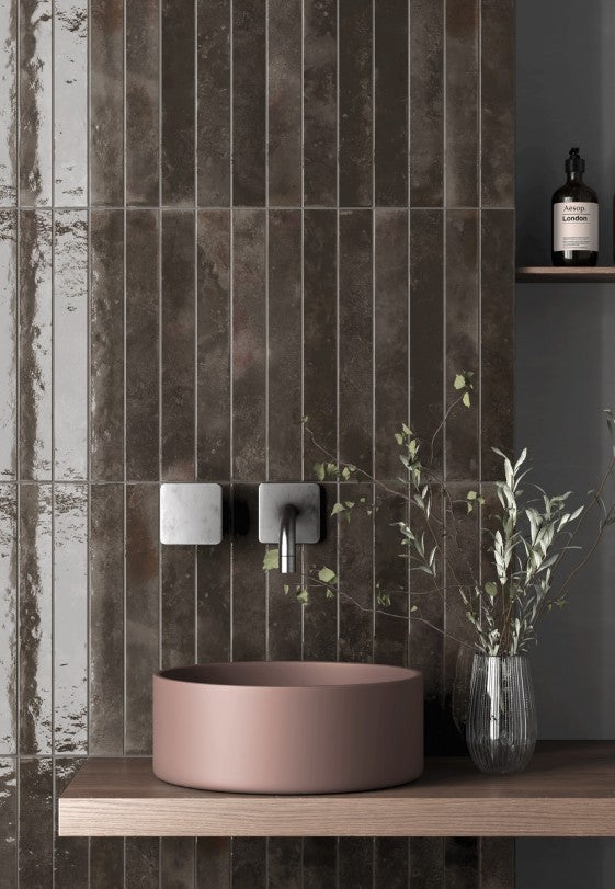 A modern bathroom scene featuring a round, matte dusty rose vessel sink mounted on a floating wooden shelf. The sink is set against a wall of dark brown textured vertical tiles, with a matching dark wood shelf below.